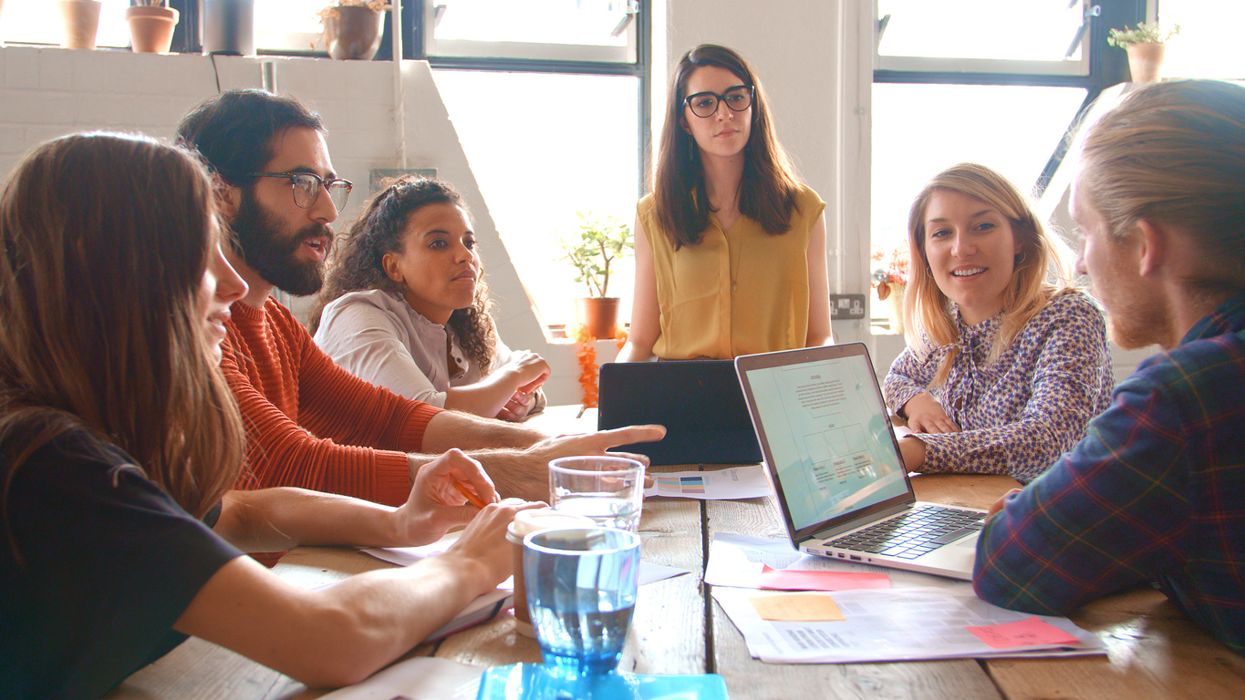 A group of six people sit around a wooden meeting table, holding a meeting. One of them has a laptop open and there's various documents scattered around a table. Each person is looking engaged and thoughtful.